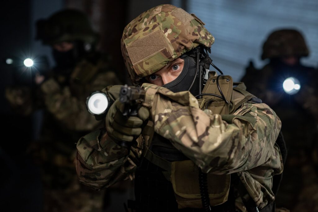 Maskierte Person in Tarnkleidung zielt bei schwachem Licht mit Pistole und Taschenlampe.